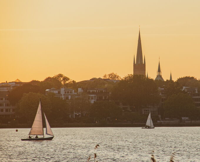 Segelboote auf der Alster vor der Hamburger Skyline im warmen Licht des Sonnenuntergangs.
