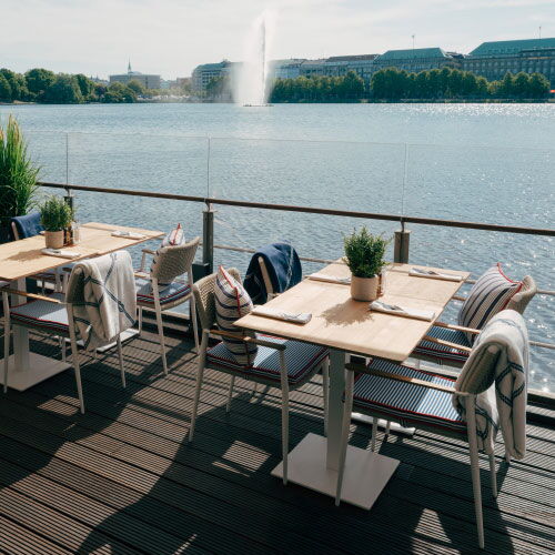 Deck 4 Terrace on the Inner Alster with wooden tables and chairs decorated with potted plants. In the distance, a fountain sprays under a clear sky.