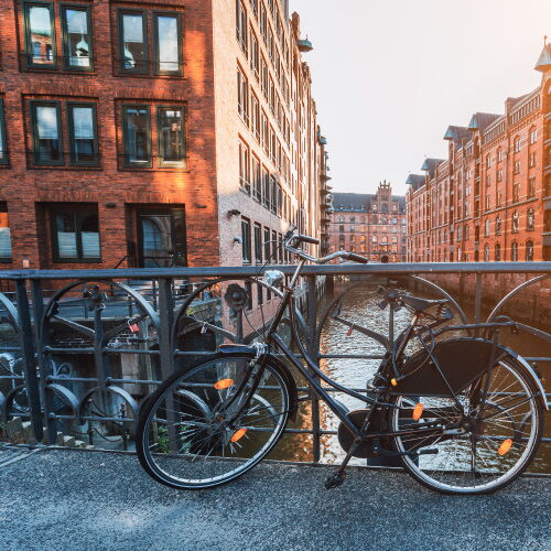 A silver bicycle is attached to a railing overlooking a canal with red brick houses. Water can be seen in the background, with a metal bridge above.