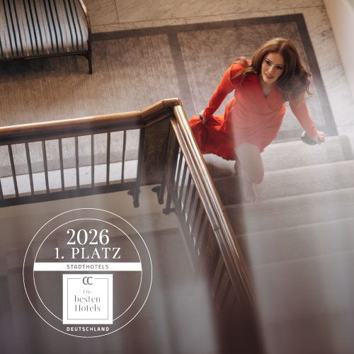 Woman in a red dress on the staircase of Hotel Vier Jahreszeiten with the “1st Place City Hotels 2026” award