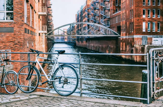 Ein silbernes Fahrrad ist an einem Geländer mit Blick auf einen Kanal mit roten Backsteinhäusern angeschlossen. Im Hintergrud ist Wasser zu sehen, darüber ist eine Metallbrücke.
