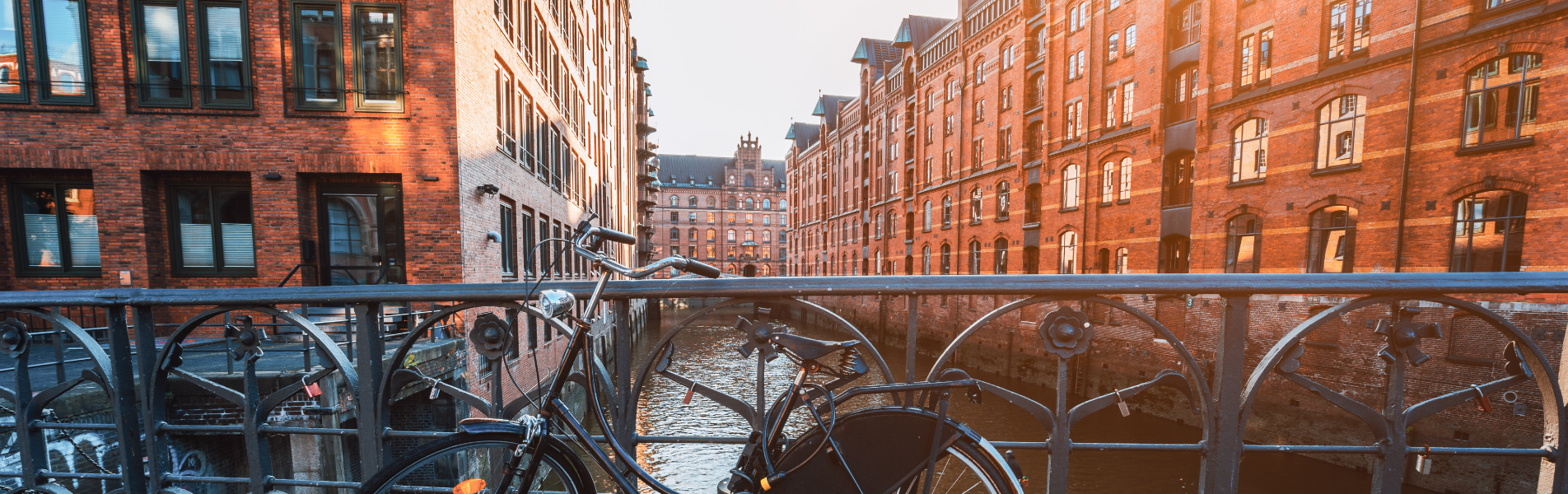 Ein silbernes Fahrrad ist an einem Geländer mit Blick auf einen Kanal mit roten Backsteinhäusern angeschlossen. Eine Metallbogenbrücke überspannt das Wasser.