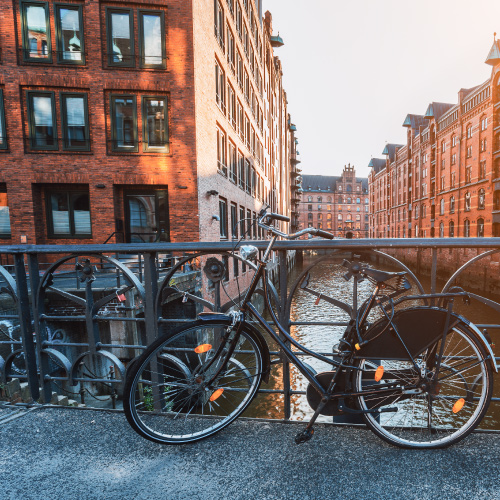 Ein silbernes Fahrrad ist an einem Geländer mit Blick auf einen Kanal mit roten Backsteinhäusern angeschlossen. Eine Metallbogenbrücke überspannt das Wasser.